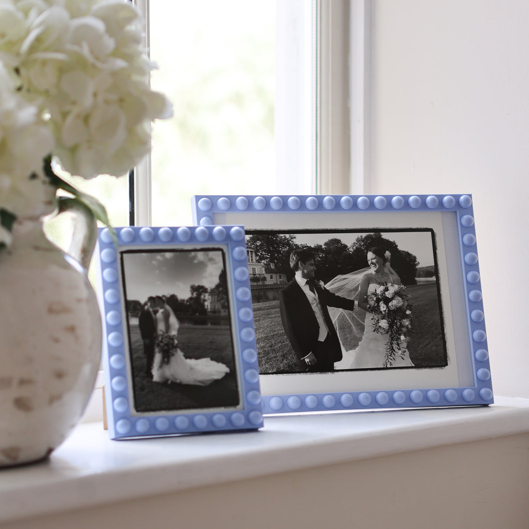 Handmade wooden bobbin frame in cornflower blue in A4 size on a windowsill with a vase full of white peonies