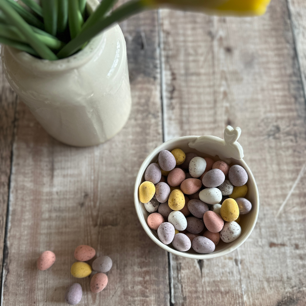 Handmade porcelain bowl filled with mini eggs on a wooden table next to a vase of daffodils